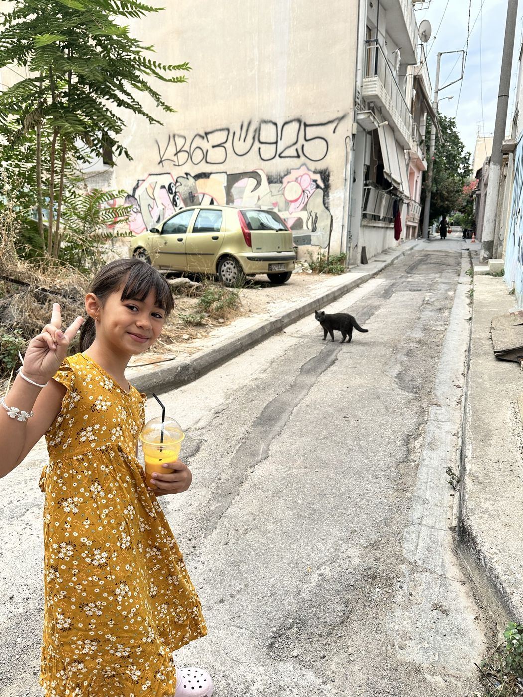 Olivia holding a fresh orange juice on a quiet Athens street, with a black cat crossing behind her