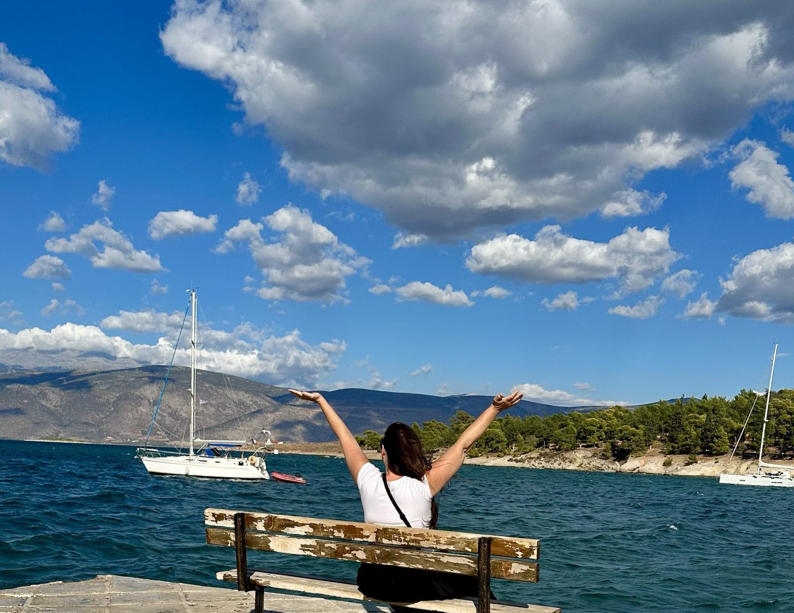 Brittany on a bench by the sea in Galaxidi, arms raised toward sailboats and mountains